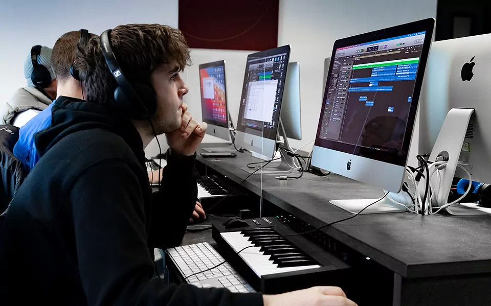 college student with headphones using an apple mac to produce music. There is a keyboard on his desk