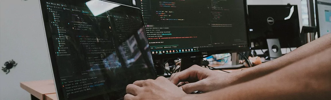 A bank of computers showing code on screen with a pair of hands coding